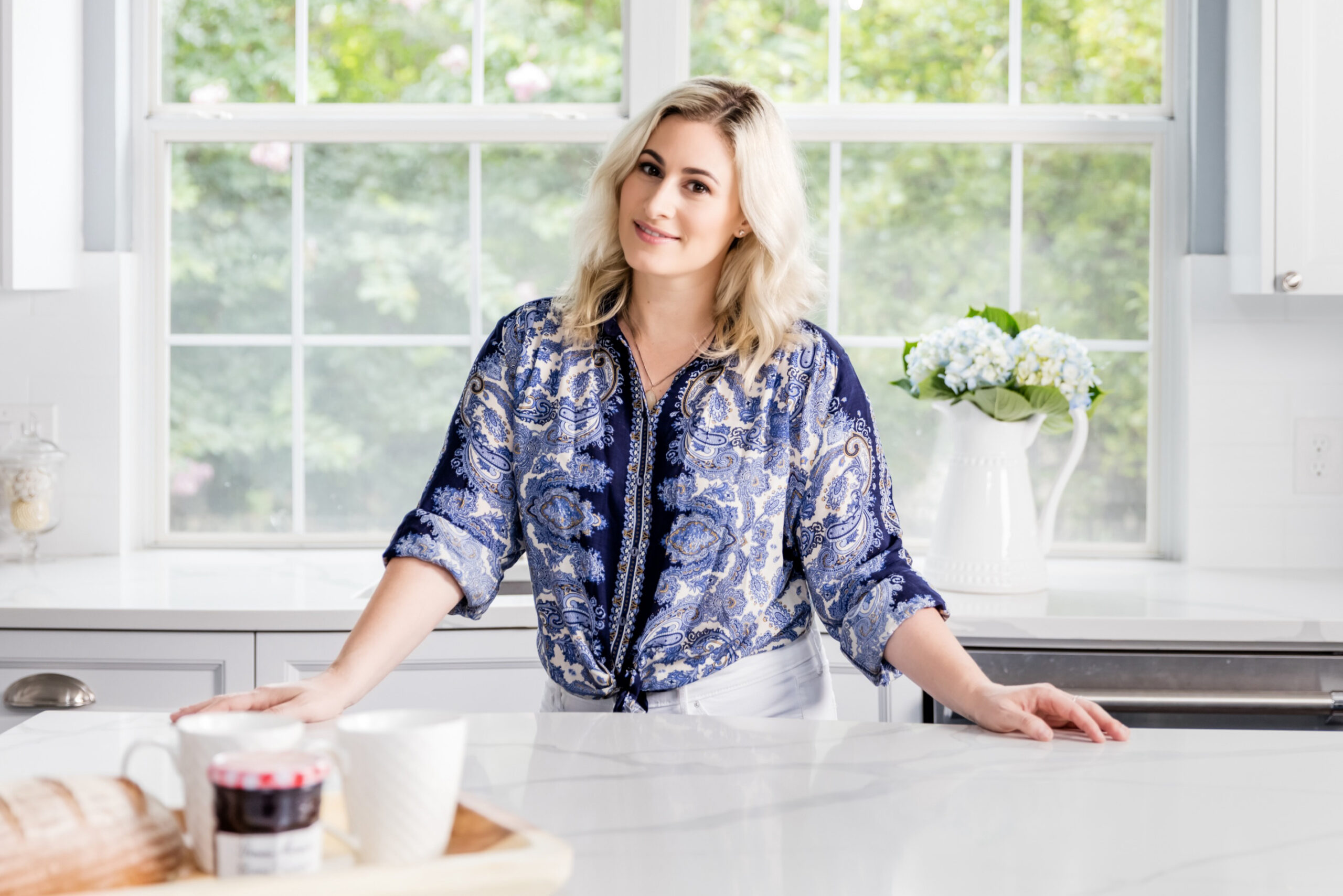 Allison Jaffe pictured in a light and airy kitchen with paned glass windows behind her overlooking a lush backyard landscape